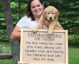 outdoor photo of woman holding a golden retriever puppy and a Gotcha Day sign