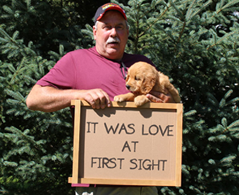 outdoor photo of man holding a golden retriever puppy and a Gotcha Day sign