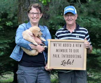 outdoor photo of couple holding a golden retriever puppy and a Gotcha Day sign