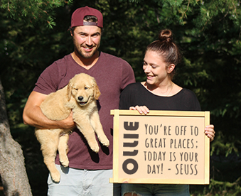 outdoor photo of a couple holding a golden retriever puppy and a Gotcha Day sign