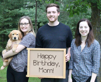 outdoor photo of two women and a man holding a golden retriever puppy and a Gotcha Day sign