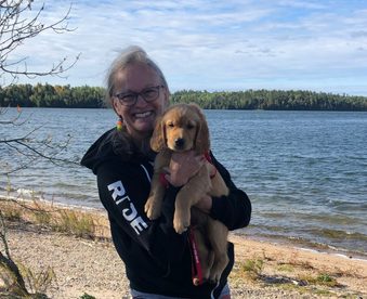 outdoor photo on lakeshore of a woman holding a golden retriever puppy and a Gotcha Day sign