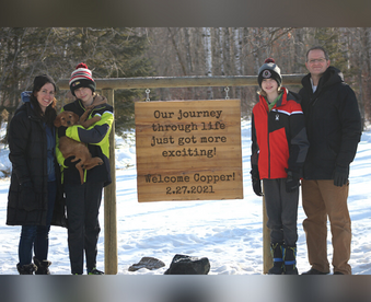 outdoor winter photo of a family of four with one member holding a golden retriever puppy and a Gotcha Day sign