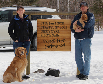 outdoor winter photo of two family members holding a golden retriever puppy and a Gotcha Day sign; adult golden retriever sitting at their feet