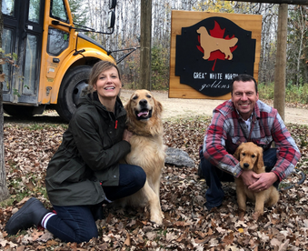 outdoor autumn photo of a couple holding a golden retriever puppy and a Gotcha Day sign, their adult golden retriever sitting with them